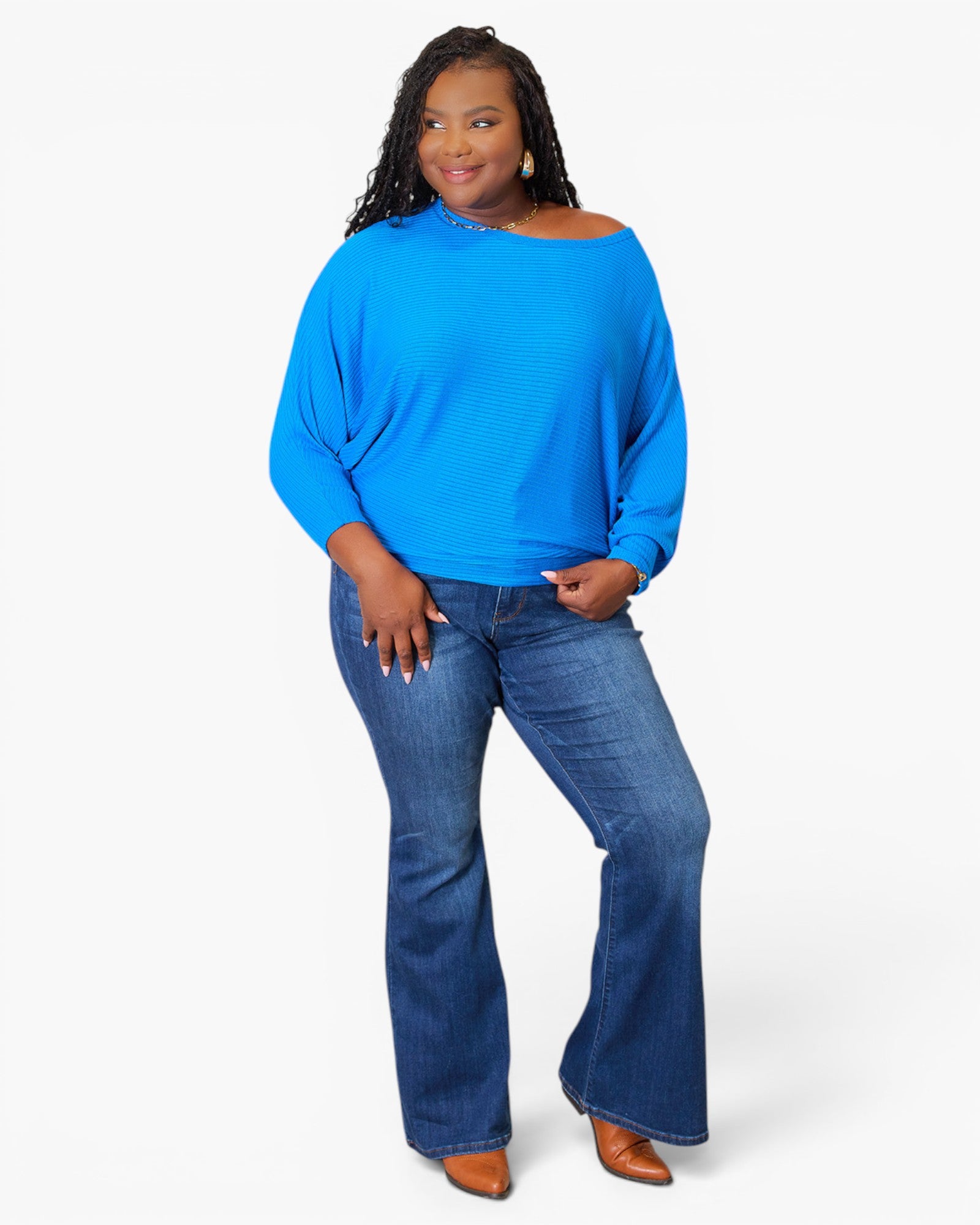 A woman modeling a bright blue off-the-shoulder long-sleeve top with blue flare jeans and brown shoes, standing against a light gray studio background.
