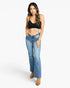 A woman modeling a pair of medium-wash blue flare jeans with a black crop top and tan ankle boots, standing against a light gray studio background.