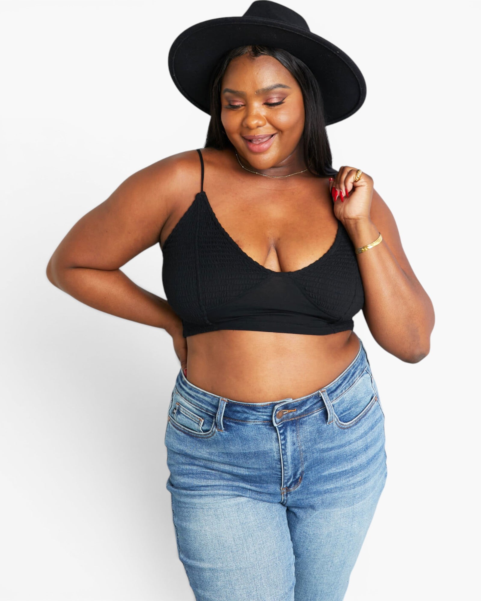 A woman modeling a black ribbed spaghetti-strap crop top with a V-neckline, paired with light-wash blue jeans and a black wide-brim hat, against a plain white studio background.