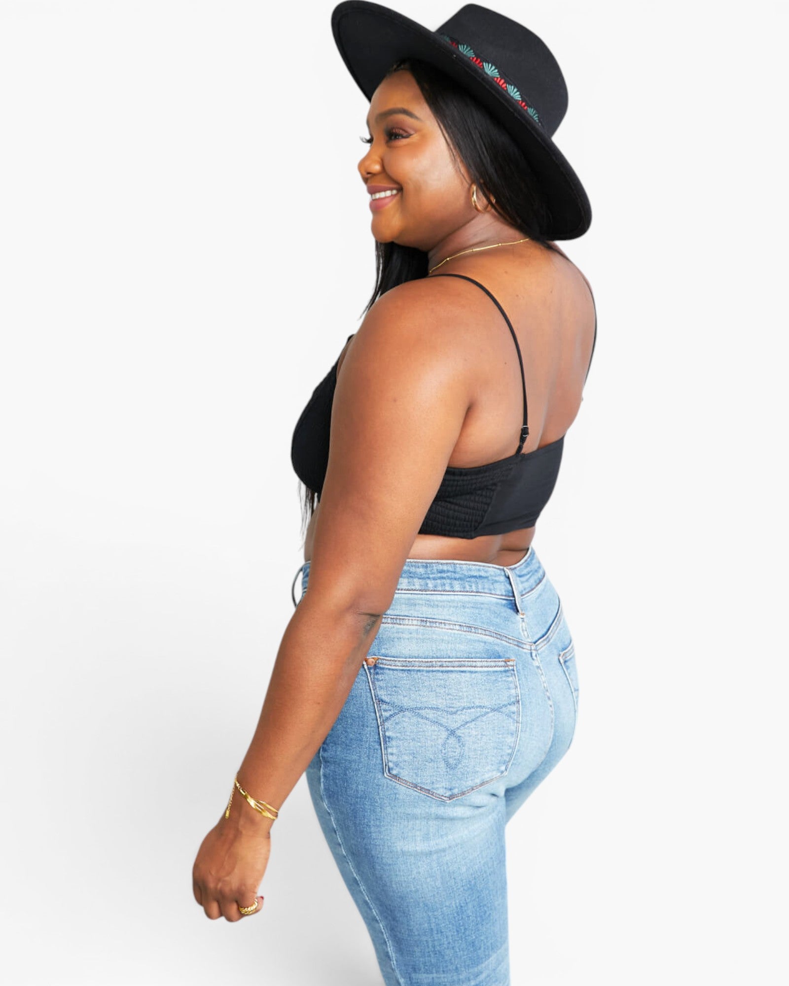 A woman wearing a black wide-brim hat with colorful trim, a black spaghetti-strap crop top, and light-wash blue jeans, shown from a side-back angle against a plain white studio background.