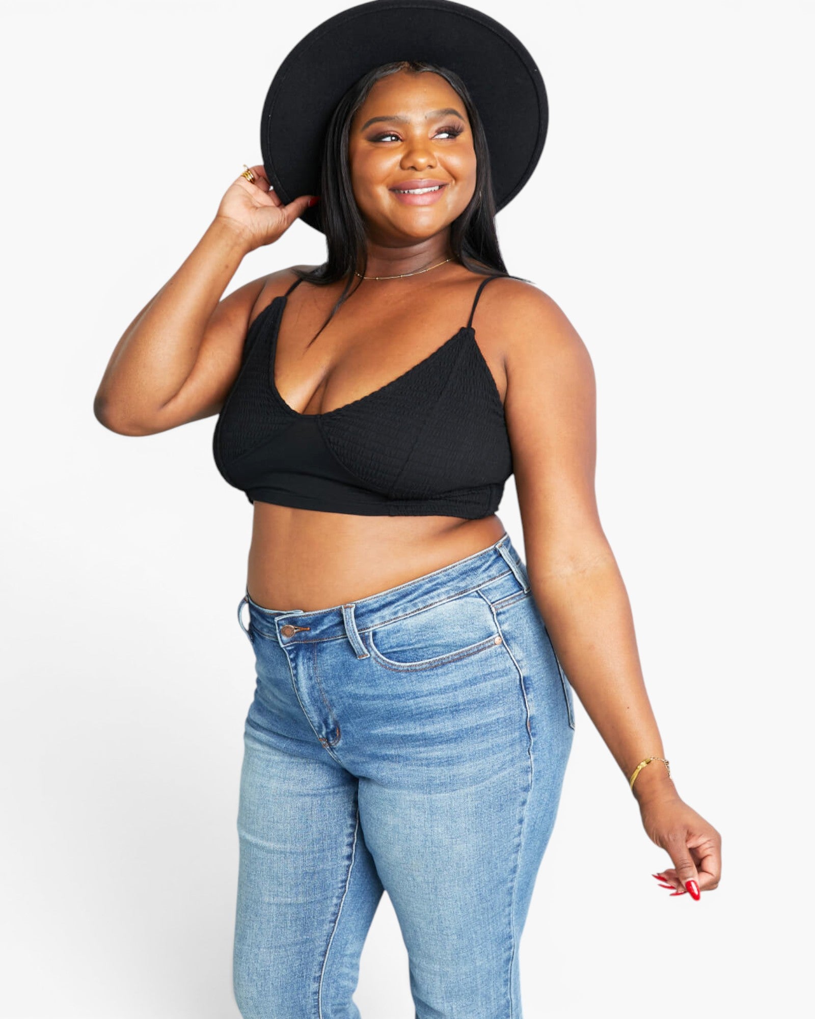A plus-size woman wearing a black ribbed spaghetti-strap crop top, blue jeans, a wide-brim black hat, and a gold bracelet, posing against a light gray studio background.
