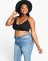 A plus-size woman wearing a black ribbed spaghetti-strap crop top, blue jeans, a wide-brim black hat, and a gold bracelet, posing against a light gray studio background.
