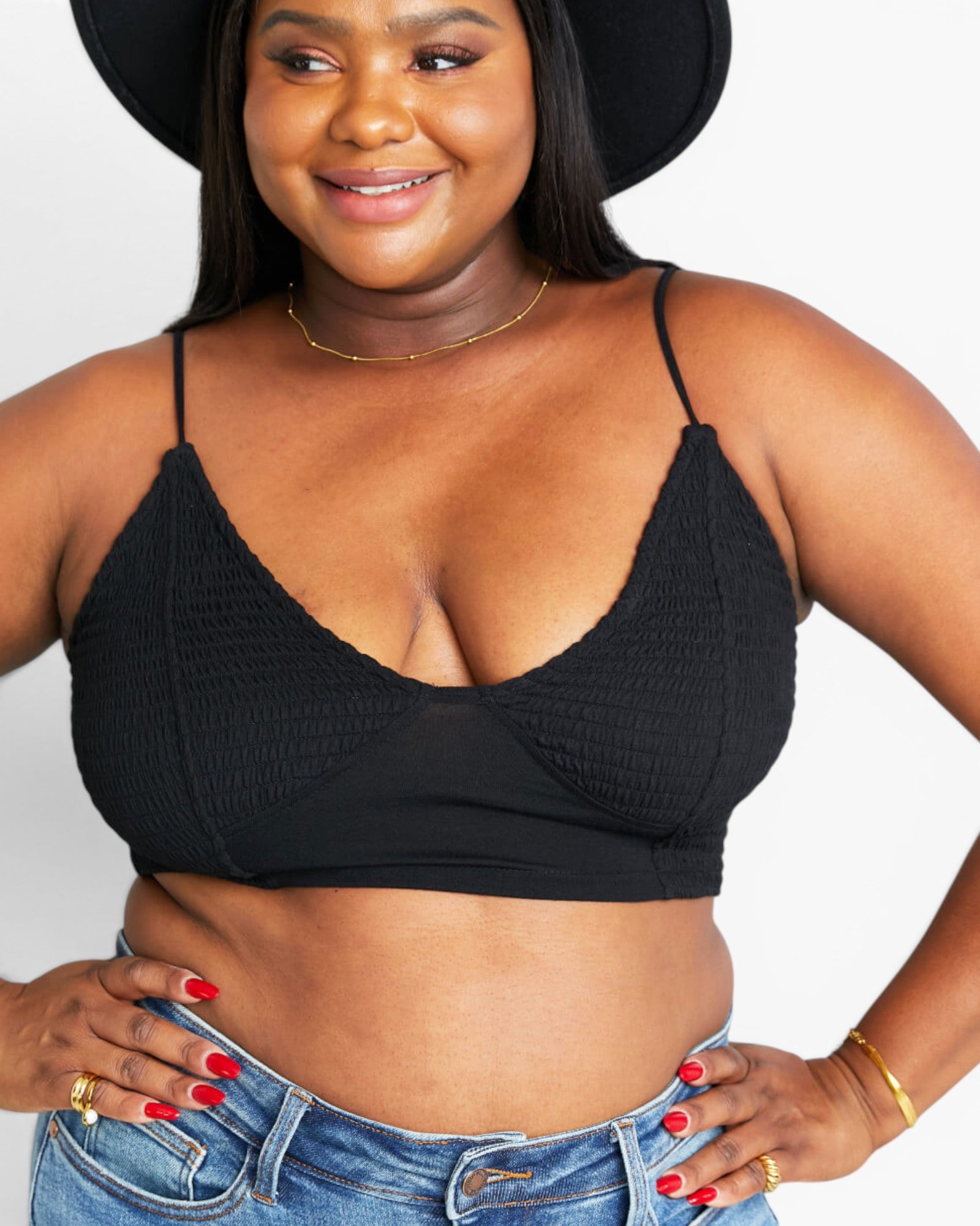 A plus-size woman wearing a black textured triangle bralette with thin straps, paired with blue jeans and a black wide-brim hat, against a plain white background.