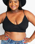 A plus-size woman wearing a black textured triangle bralette with thin straps, paired with blue jeans and a black wide-brim hat, against a plain white background.