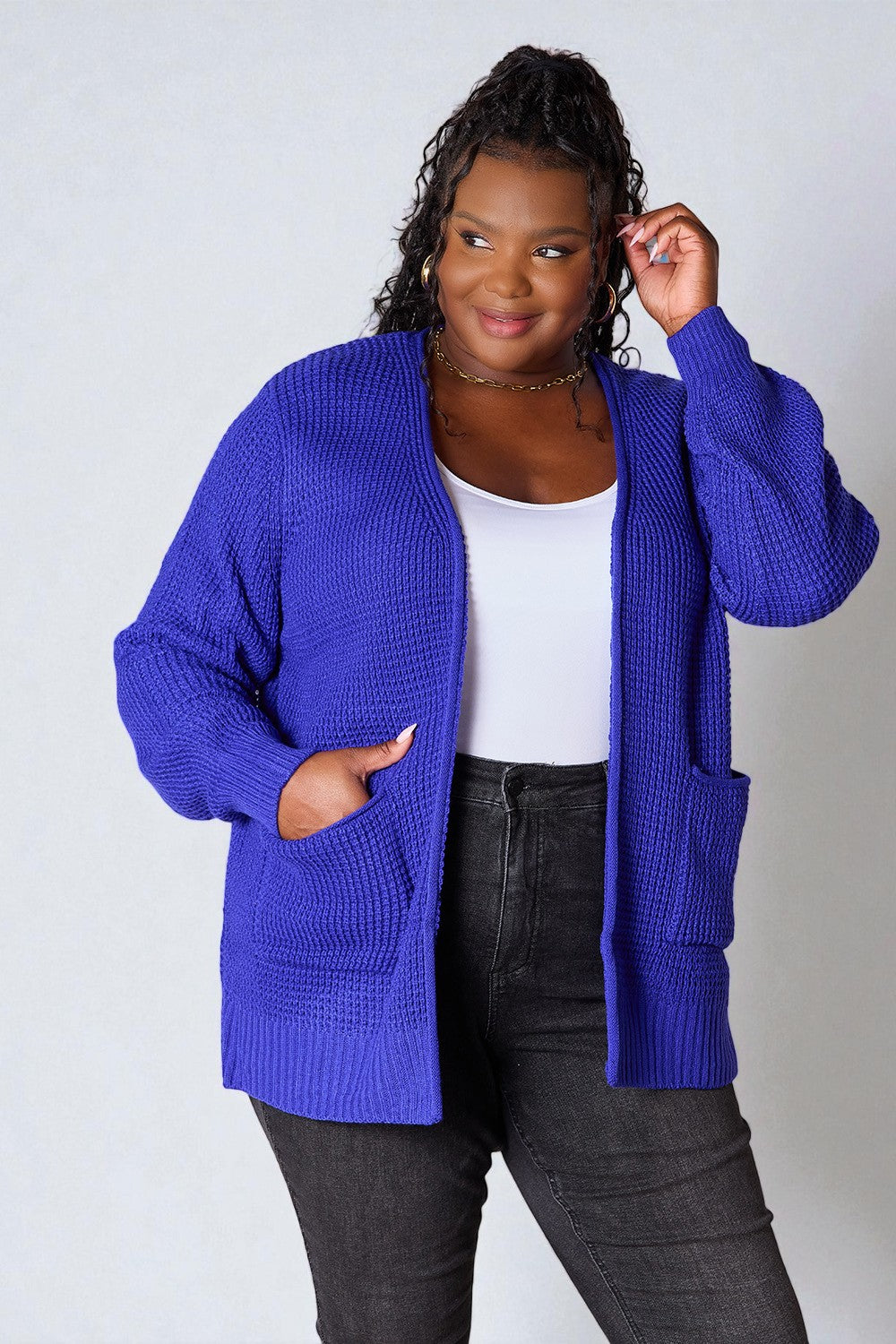 Woman wearing a blue cardigan over a white top and black pants on a plain background