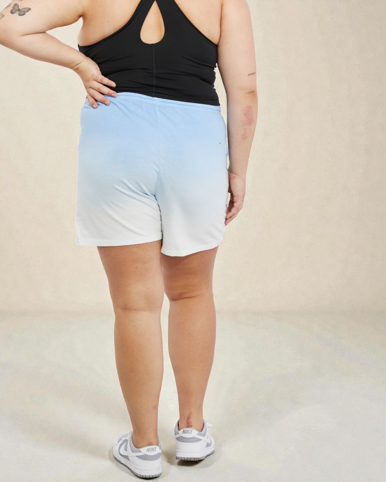 Person wearing a black tank top and light blue to white ombre shorts on a beige background
