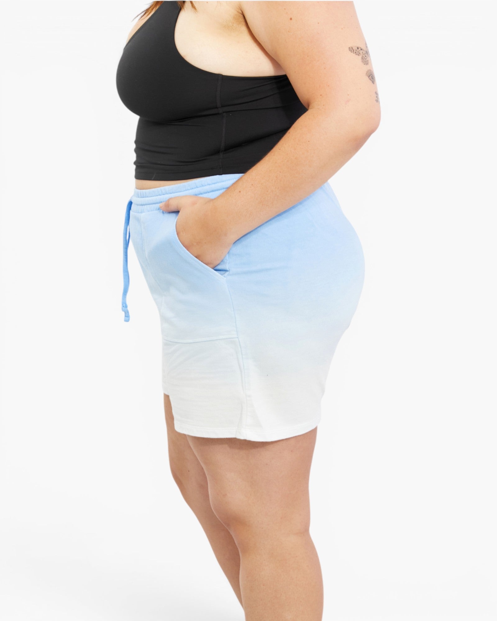 A side-profile view of a model wearing a light blue and white ombre mini skirt with a drawstring waist and side pockets, paired with a black tank top, against a plain white background.