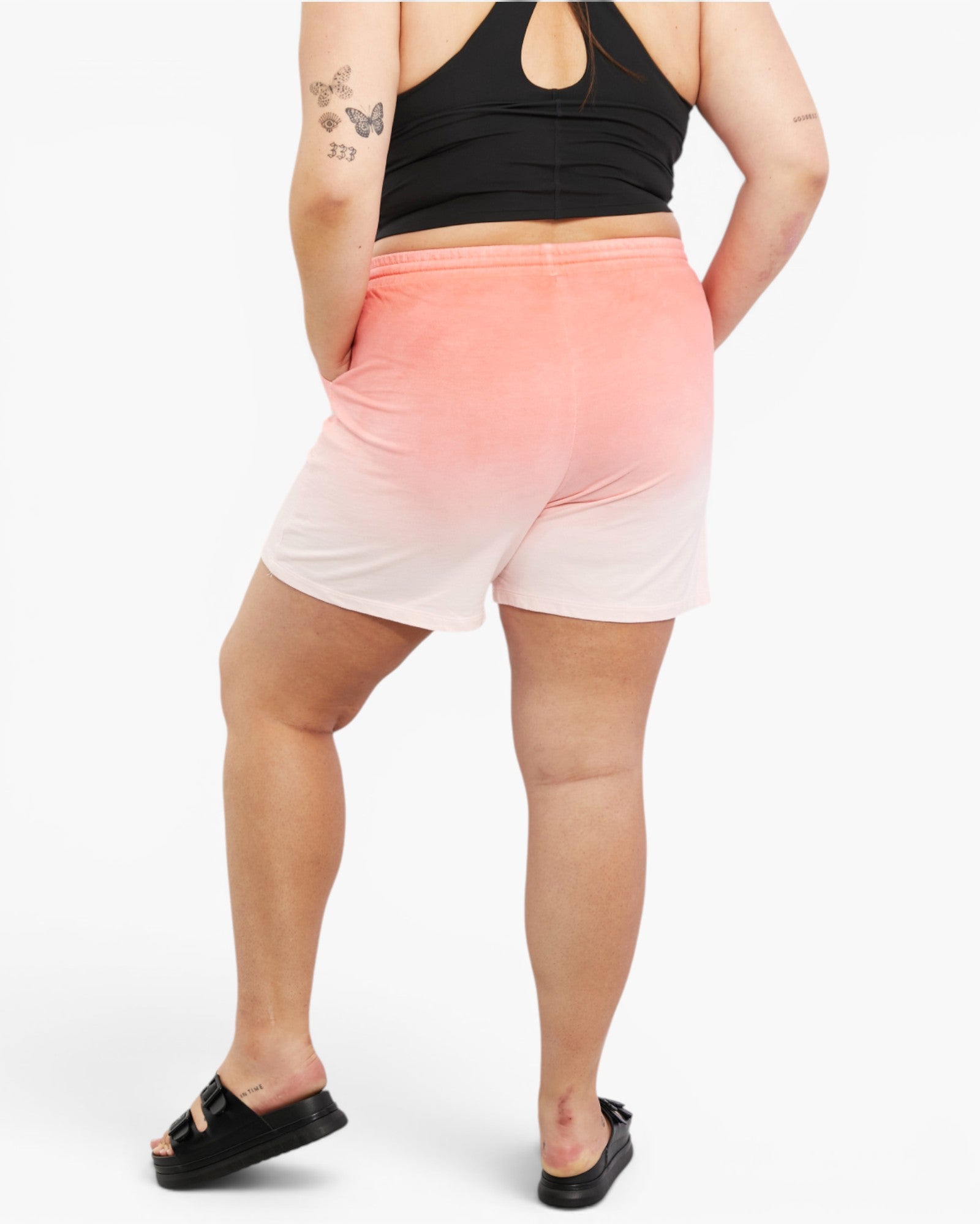 A person shown from the back wearing gradient athletic shorts that fade from coral pink at the top to white at the bottom, paired with a black sports bra and black slide sandals on a light gray studio background.