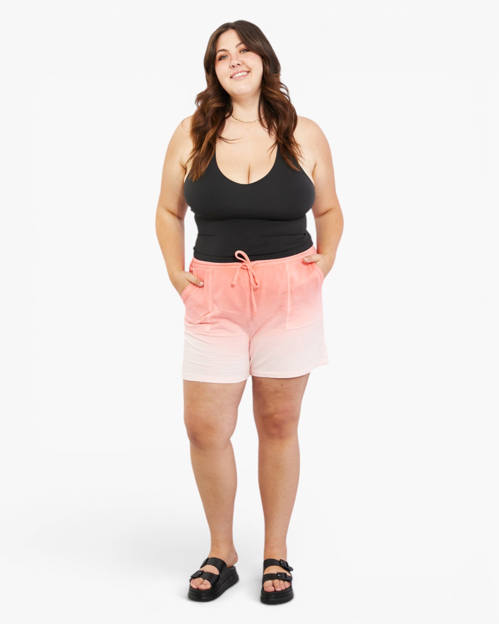 A woman modeling a pair of pink and white ombre gradient drawstring shorts, paired with a black tank top and black slide sandals, against a plain white studio background.
