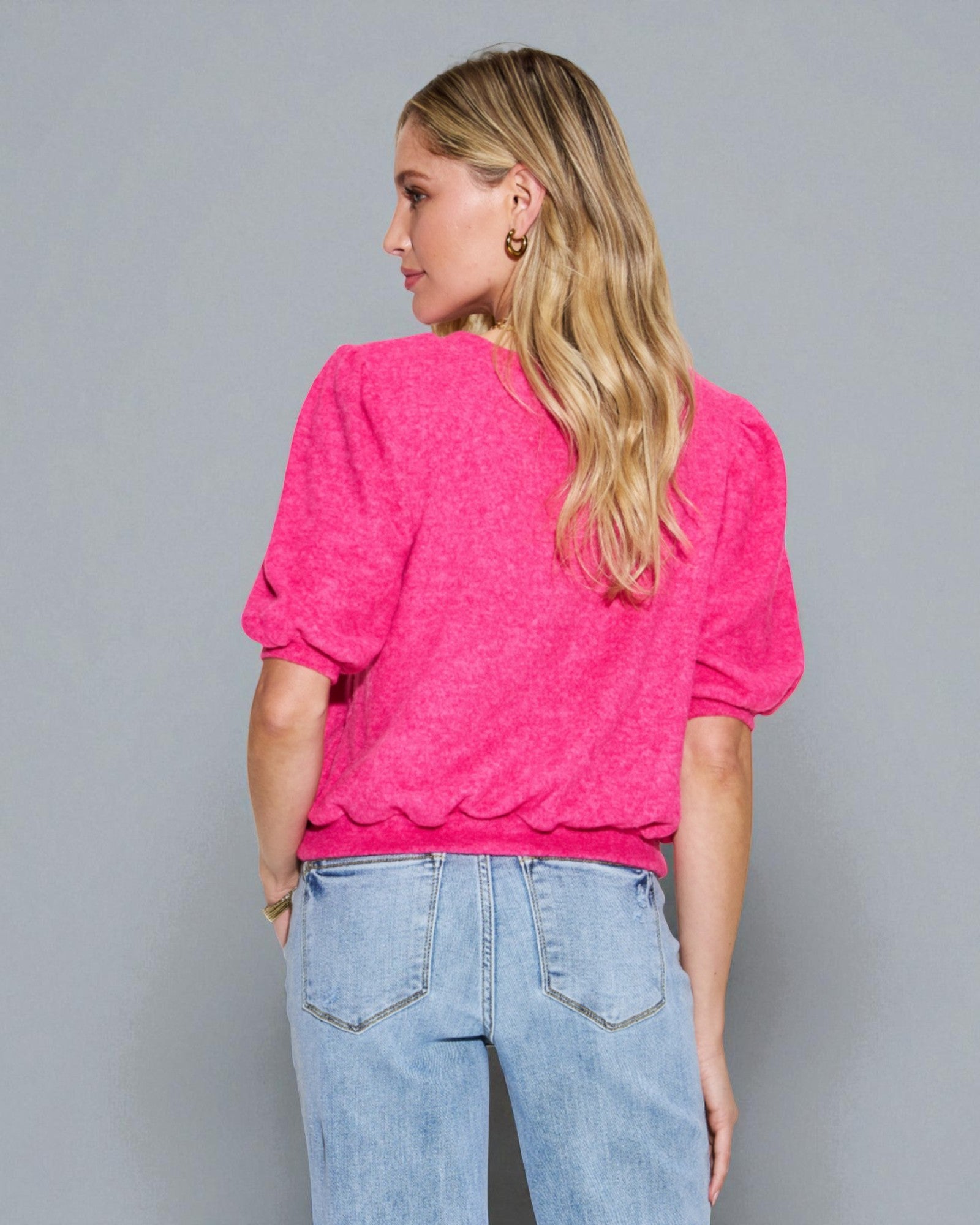 Woman wearing a bright pink top with puff sleeves and light blue jeans against a gray background