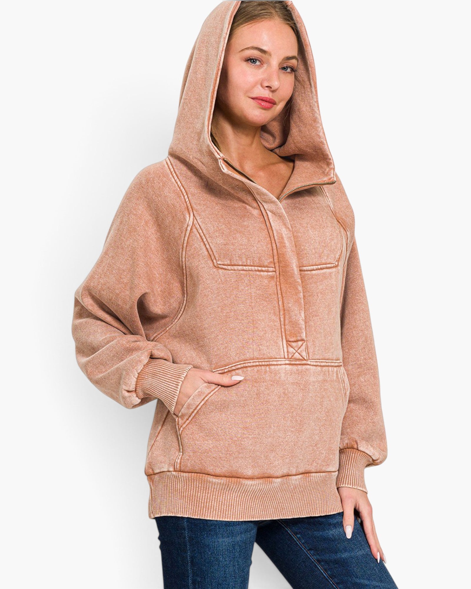 A woman wearing a faded dusty rose/peach pullover hoodie with a half-zip front, ribbed cuffs and hem, and a kangaroo pocket, paired with blue jeans against a light gray studio background.