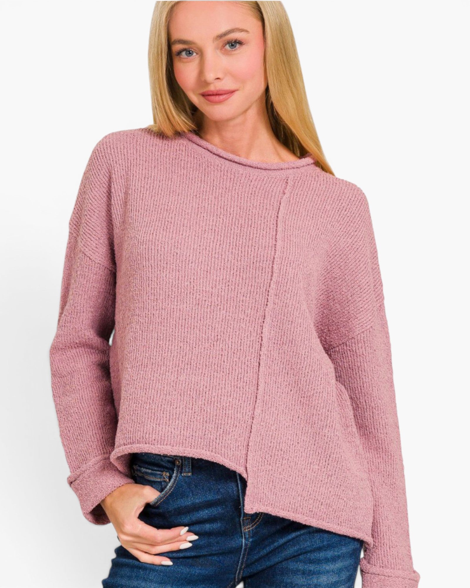 A woman wearing a dusty pink ribbed knit sweater with an asymmetrical front panel and blue jeans, photographed against a plain light background.