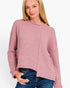 A woman wearing a dusty pink ribbed knit sweater with an asymmetrical front panel and blue jeans, photographed against a plain light background.