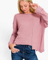 A woman wearing a dusty pink ribbed knit sweater with a relaxed fit and blue jeans, photographed against a plain white studio background.