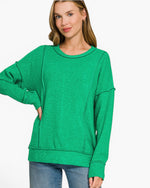 A woman wearing a bright green heathered long-sleeve sweatshirt with a relaxed, oversized fit and side pockets, paired with blue jeans, against a plain white background.