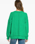 A woman viewed from the back wearing a bright green oversized knit sweater with long sleeves and a relaxed fit, paired with blue jeans against a plain white studio background.
