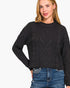 A woman wearing a black cable-knit cropped sweater with blue jeans, photographed against a plain white studio background.
