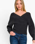 A woman wearing a black long-sleeve knit top with a wide V-neck and wrap-style crossover front, paired with blue jeans, against a plain light background.