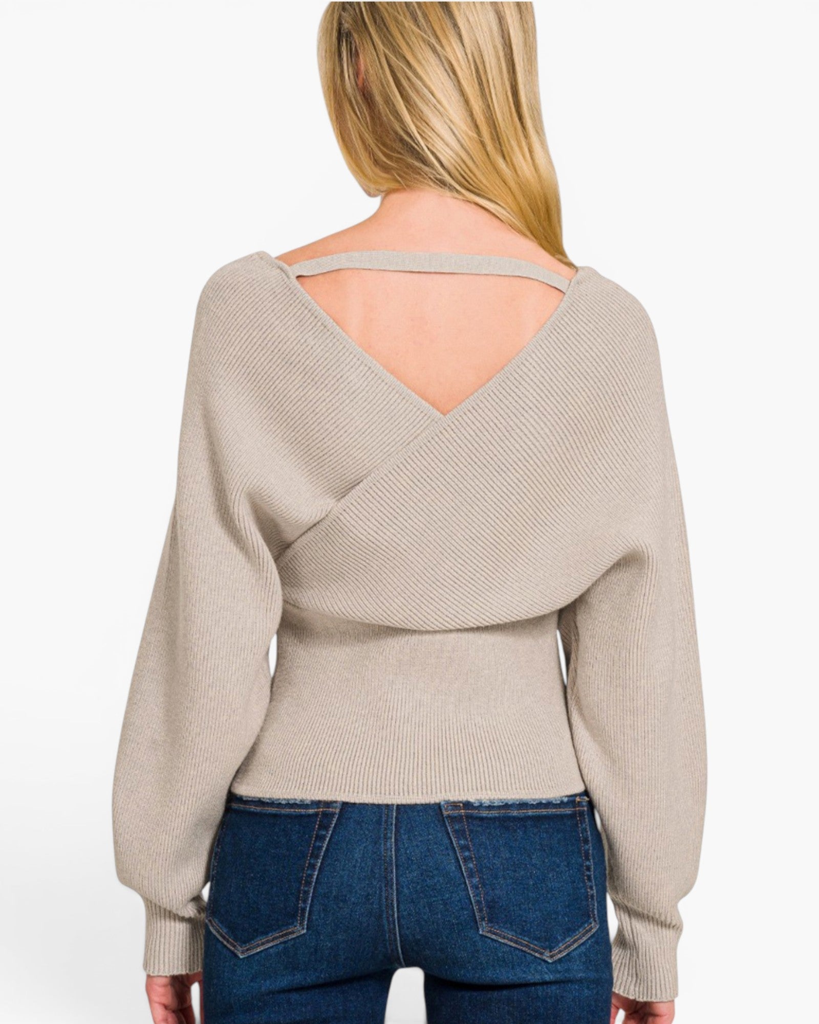 A woman shown from the back wearing a light beige ribbed knit sweater with a crossover wrap-style back and a thin shoulder strap, paired with blue jeans against a plain white background.