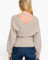 A woman shown from the back wearing a light beige ribbed knit sweater with a crossover wrap-style back and a thin shoulder strap, paired with blue jeans against a plain white background.