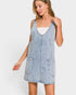 A woman wearing a light-wash denim overall-style dress over a white tank top, photographed against a plain light gray studio background.