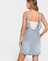 A woman shown from the back wearing a light-wash denim overall dress over a white tank top, photographed against a plain light gray studio background.