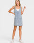 A woman modeling a light-wash denim overall mini dress over a white tank top, paired with white sneakers, against a plain light gray studio background.