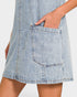 Close-up of a light blue denim dress with a large front patch pocket and a hand tucked into the pocket, shown against a plain light background.