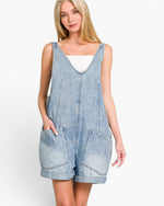 A woman modeling a light blue denim overall romper with a scoop neckline, front pockets, and rolled cuffs, worn over a white top against a plain white background.