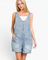 A woman modeling a light blue denim overall romper with a scoop neckline, front pockets, and rolled cuffs, worn over a white top against a plain white background.