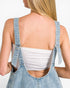 Back view of a woman wearing light blue denim overalls over a white bandeau top, showing the open back design and adjustable shoulder straps.