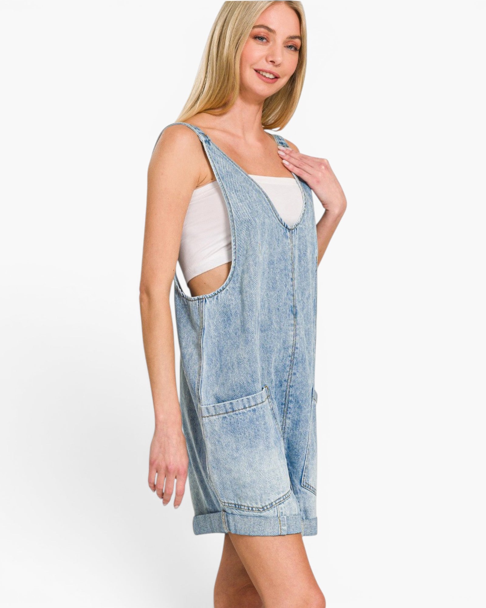 A woman modeling a light-wash denim overall mini dress over a white tank top, photographed against a clean white studio background.