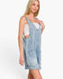 A woman modeling a light-wash denim overall mini dress over a white tank top, photographed against a clean white studio background.