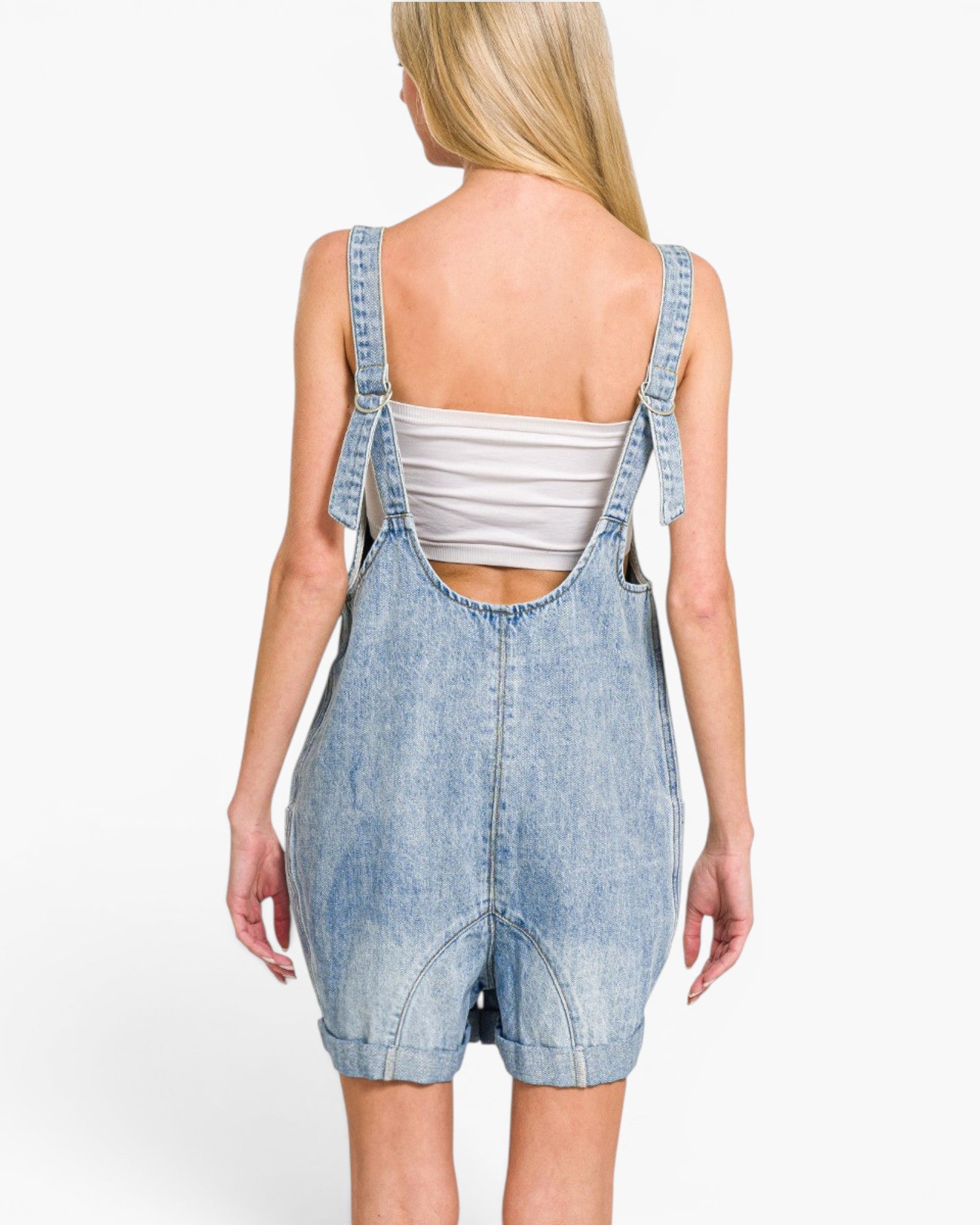 A woman shown from the back wearing a light blue denim overall romper over a white ribbed tank top, against a plain white studio background.