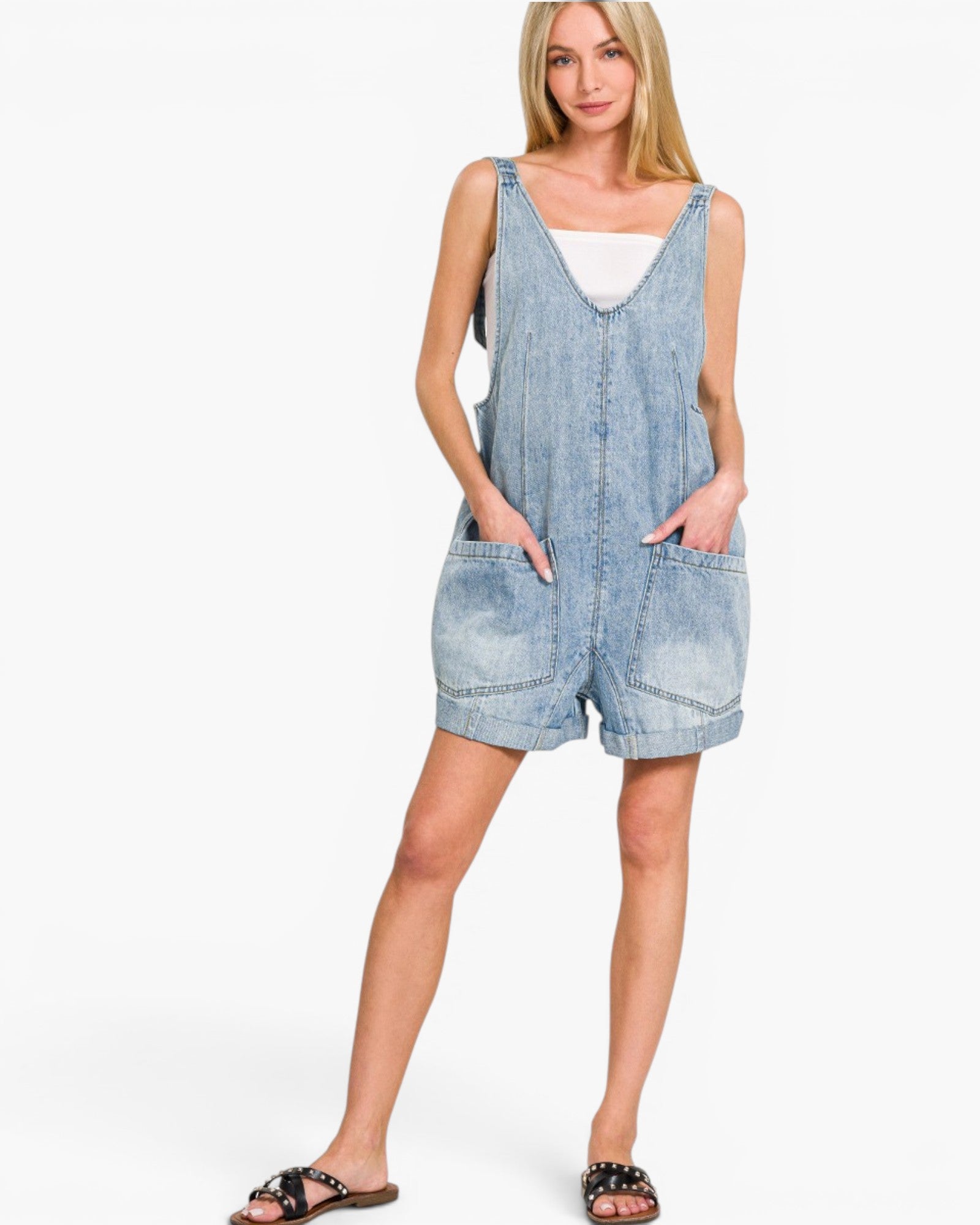 A woman modeling a light-wash denim overall romper with front pockets, worn over a white tank top and styled with black slide sandals, against a plain white studio background.