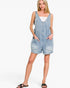 A woman modeling a light-wash denim overall romper with front pockets, worn over a white tank top and styled with black slide sandals, against a plain white studio background.