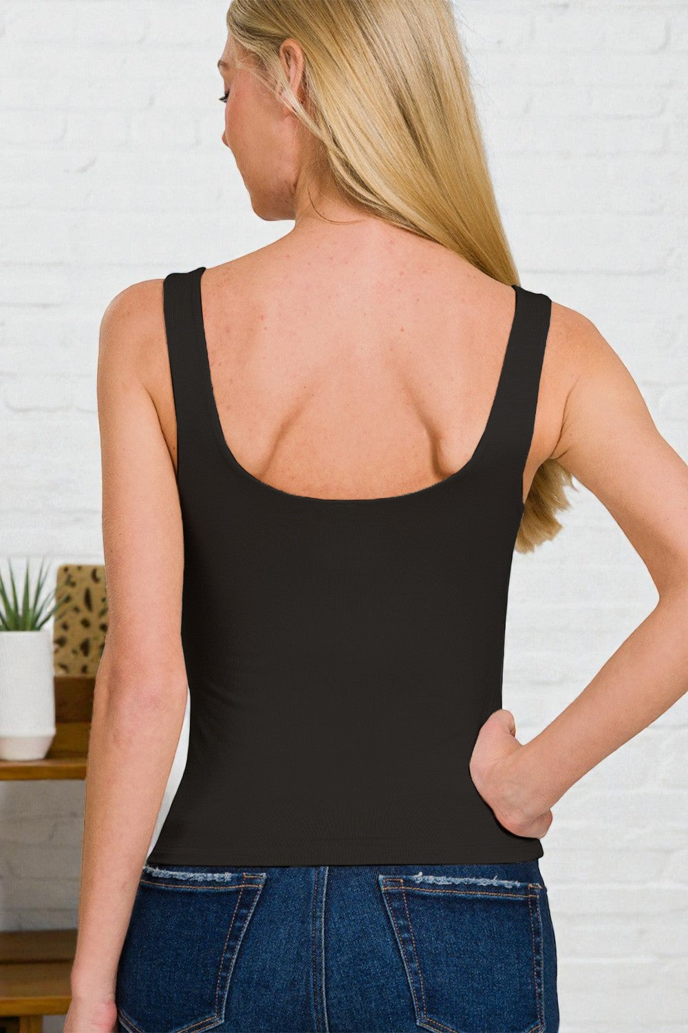 Woman wearing a black tank top and blue jeans against a white brick wall.
