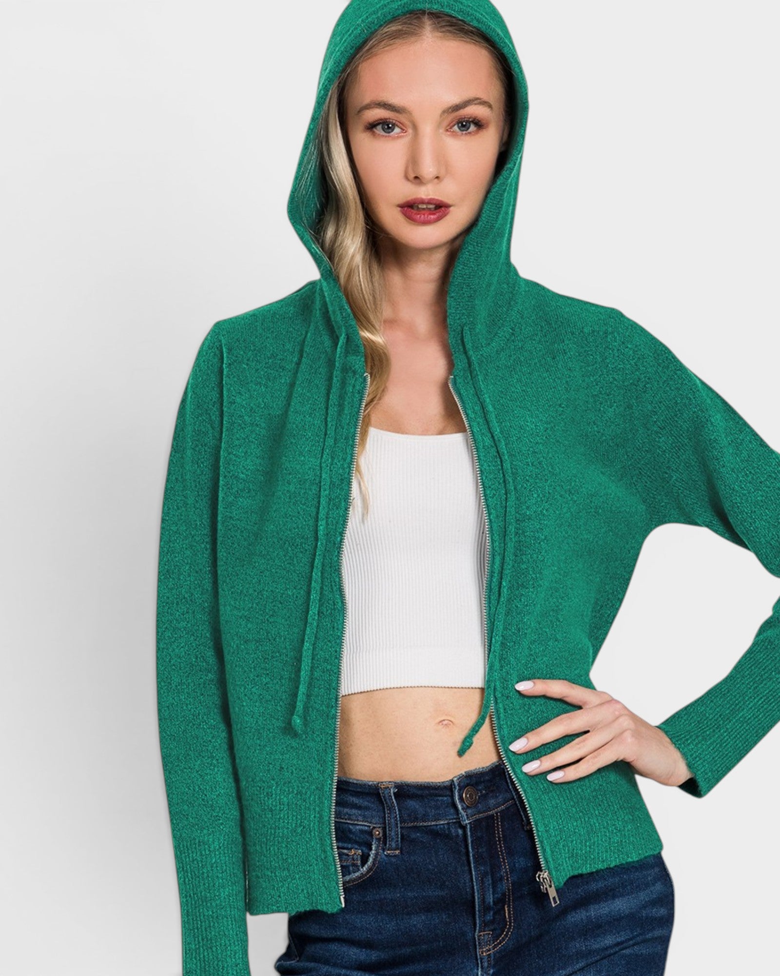 Woman wearing a green hoodie over a white top and blue jeans on a light gray background