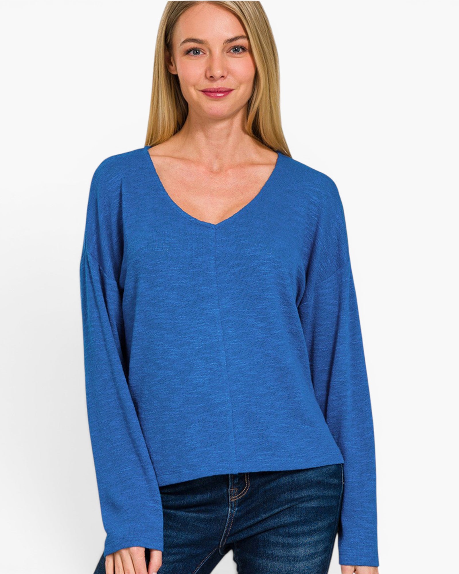 A woman wearing a blue long-sleeve V-neck top and dark blue jeans, standing against a plain light gray studio background.