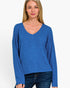 A woman wearing a blue long-sleeve V-neck top and dark blue jeans, standing against a plain light gray studio background.