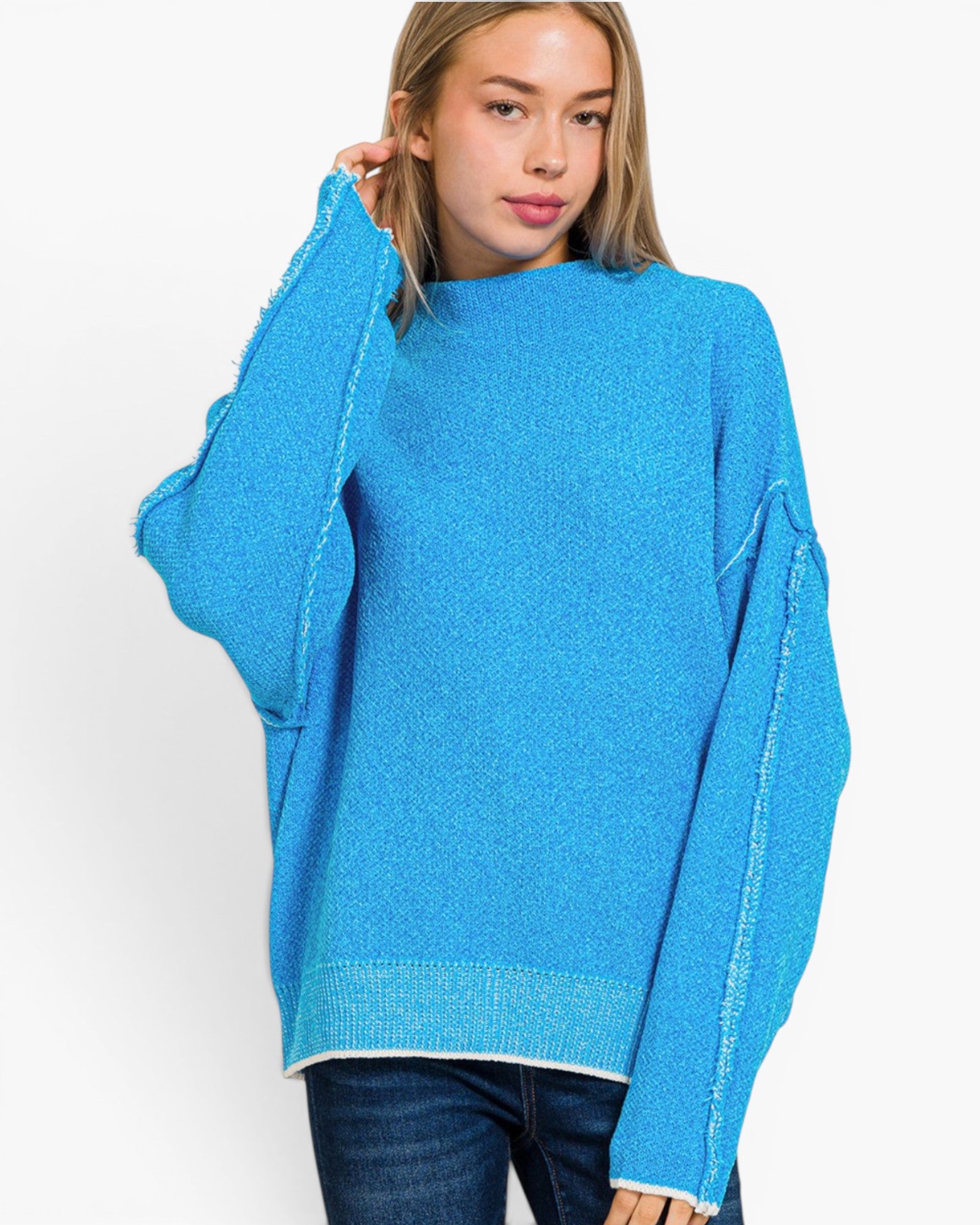 A woman wearing a bright blue knit sweater with textured, frayed edges and oversized sleeves, paired with dark blue jeans, against a plain white studio background.