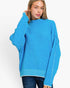 A woman wearing a bright blue knit sweater with textured, frayed edges and oversized sleeves, paired with dark blue jeans, against a plain white studio background.