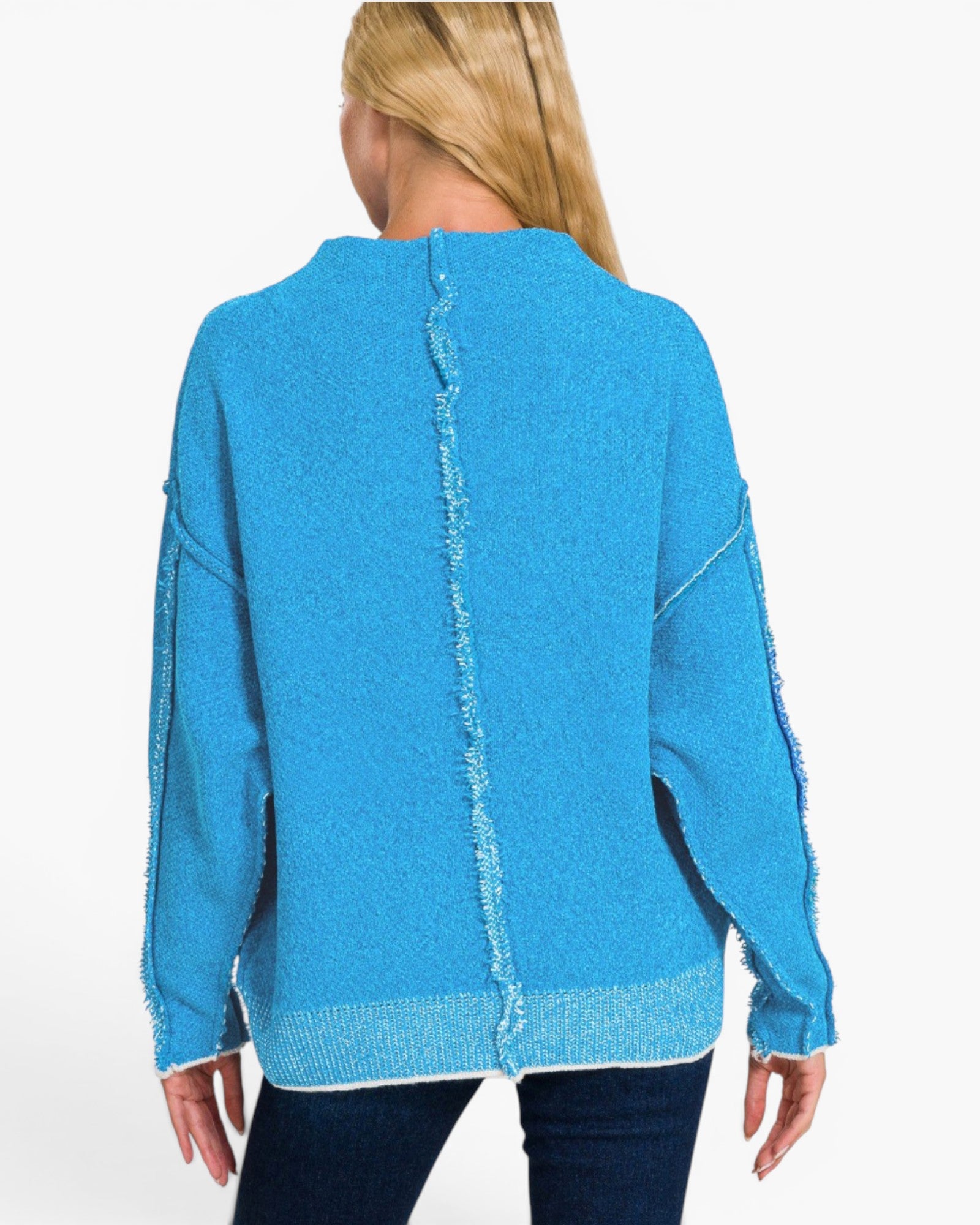 A woman viewed from the back wearing a bright turquoise blue knit sweater with a central fringe detail and frayed trim along the sleeves and hem, paired with dark blue jeans against a plain white background.