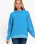 A woman wearing a bright turquoise blue knit sweater with textured, frayed sleeve details and a ribbed hem, paired with dark blue jeans, against a plain white background.