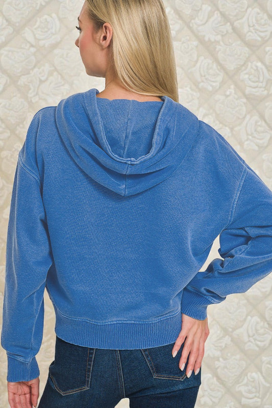 Person wearing a blue hoodie and jeans against a textured white wall.