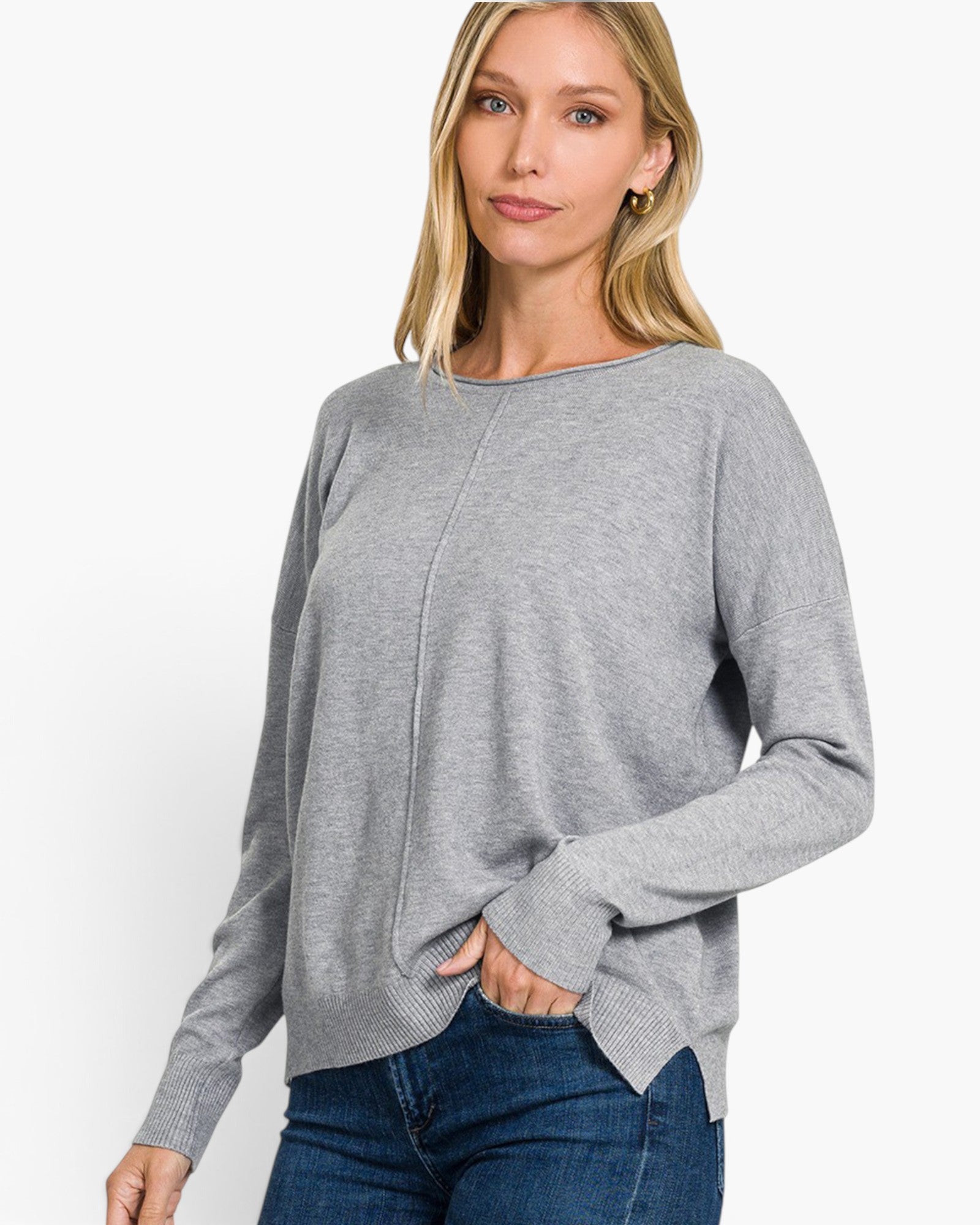 A woman wearing a light gray knit sweater with a boat neckline and long sleeves, paired with blue jeans, photographed against a clean white studio background.