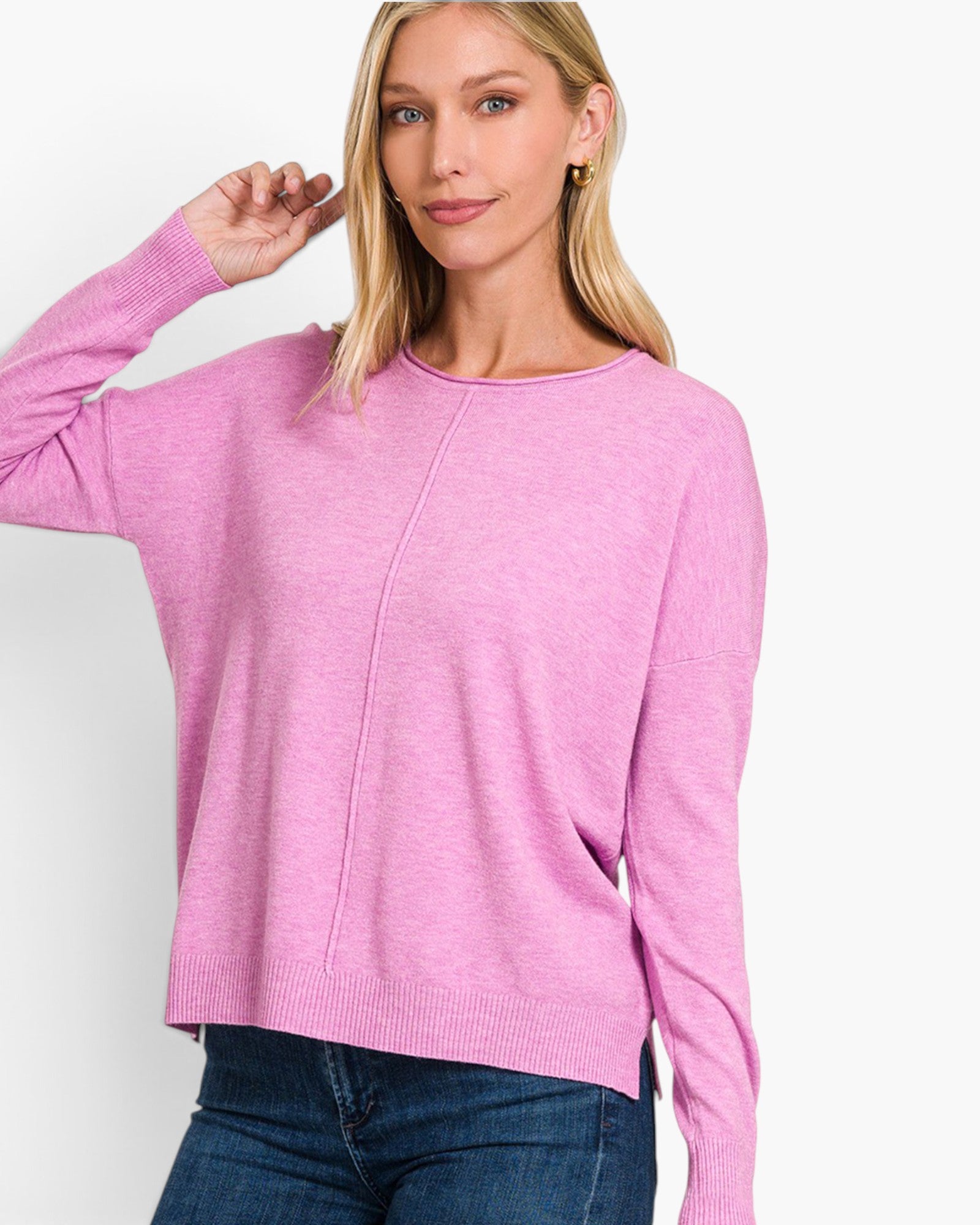 A woman wearing a pink long-sleeve knit sweater with a crew neck and front seam detail, paired with blue jeans, against a plain white studio background.