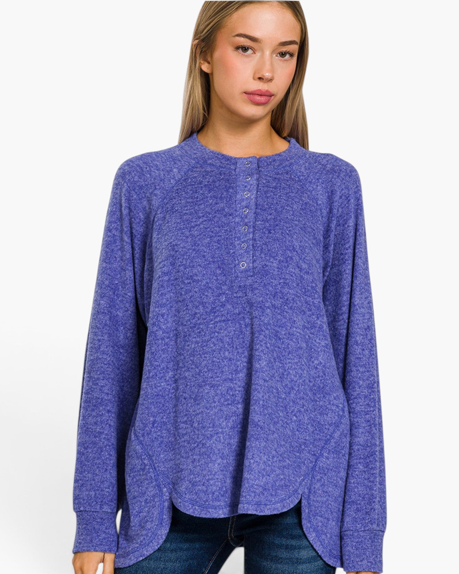 A woman wearing a heathered periwinkle blue long-sleeve henley top with a buttoned neckline, paired with dark blue jeans, against a plain white background.