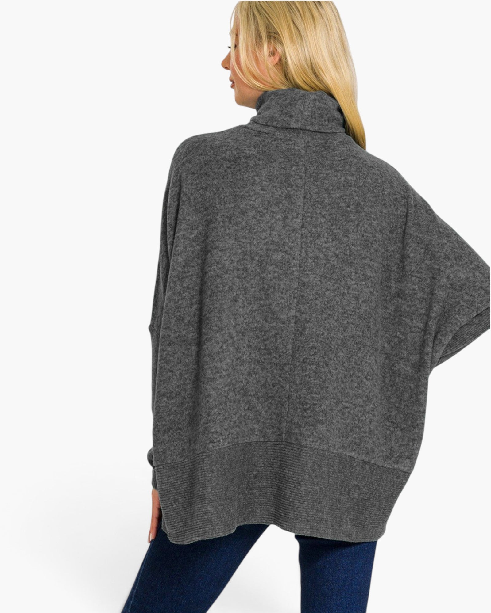 A woman shown from the back wearing a dark gray heathered knit poncho sweater with a high folded collar and wide draped sleeves, paired with blue jeans, against a plain white studio background.