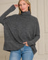 Woman wearing a gray turtleneck sweater and blue jeans against a neutral wall.